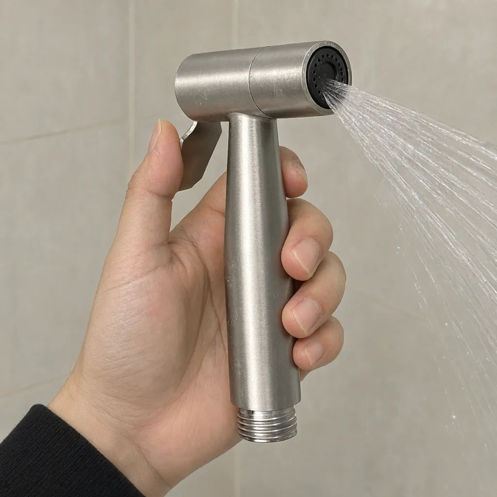 Hand holding a brushed silver handheld bidet sprayer with water spraying from the nozzle