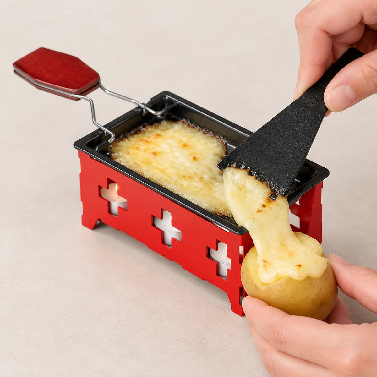 Hands scraping melted raclette cheese from a red mini raclette set onto a potato