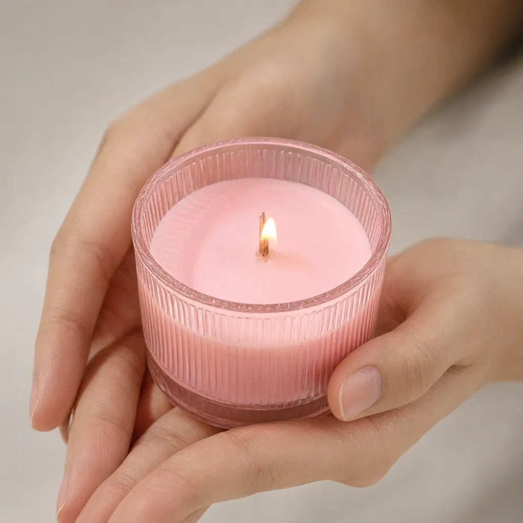 Hands holding a lit pink scented candle in a ribbed glass jar with a wooden wick