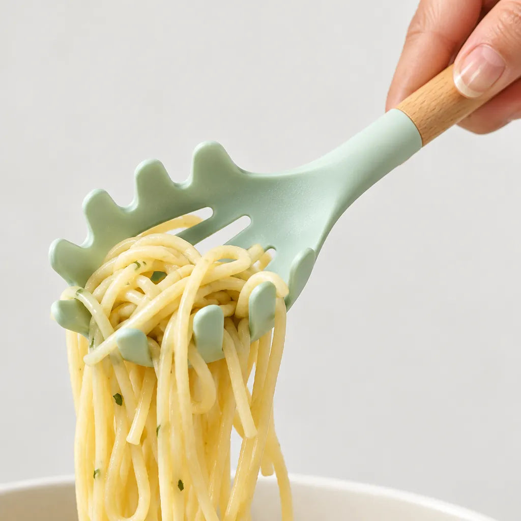 Hand lifting pasta with a mint green silicone pasta server with wooden handle from a kitchen utensil set