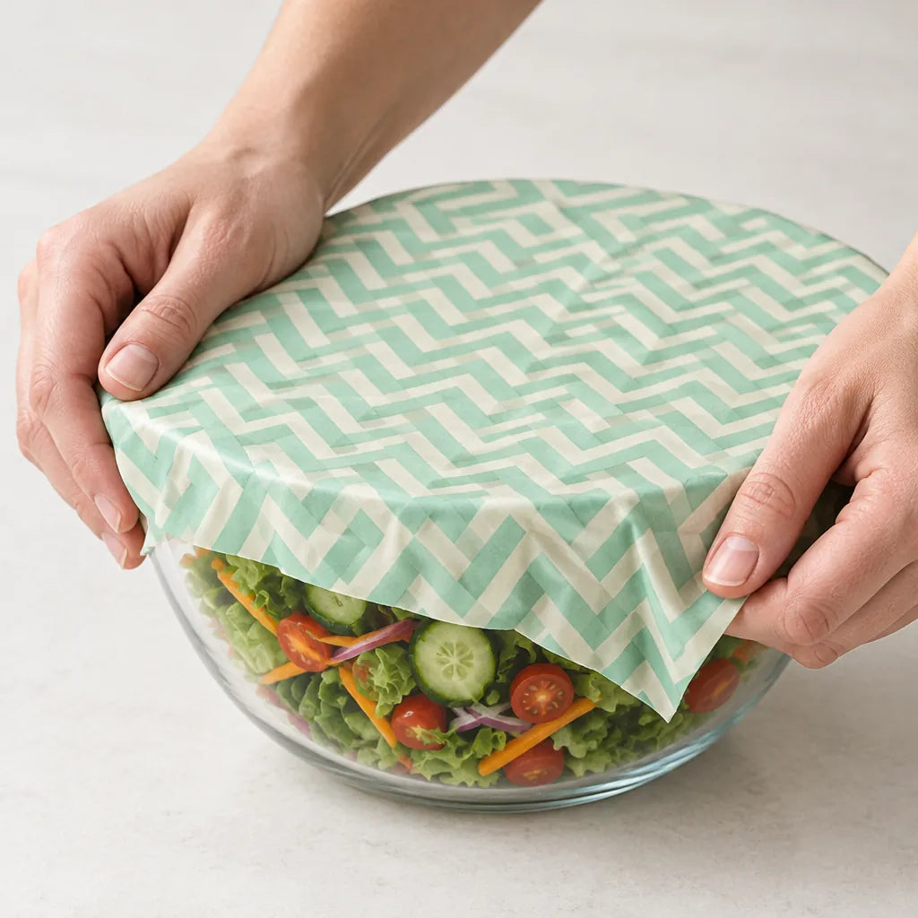 Hands pressing a mint chevron beeswax wrap over a glass bowl of salad