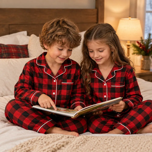 Two kids in matching plaid Christmas pajamas reading a book on a bed