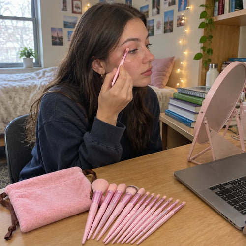 Studentin am Schreibtisch trägt Lidschatten mit einem kleinen rosa Schminkpinsel auf