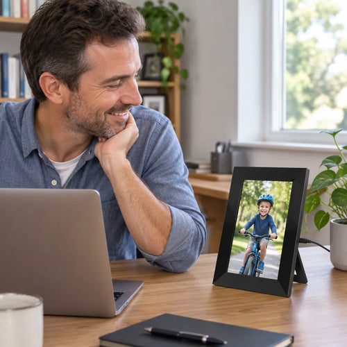 Man at his home office desk smiling at a digital frame standing upright with a photo of his son