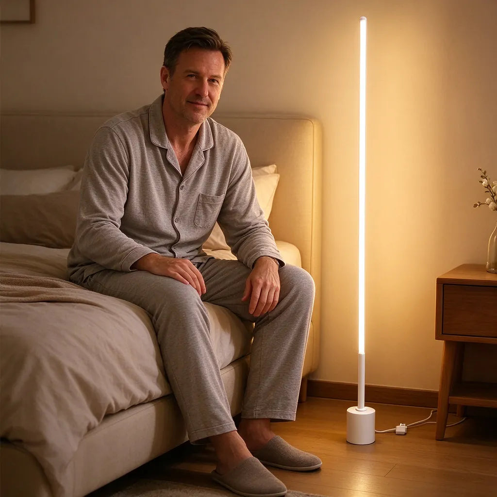 Man sitting on a bed beside a slim white LED floor lamp glowing warm light