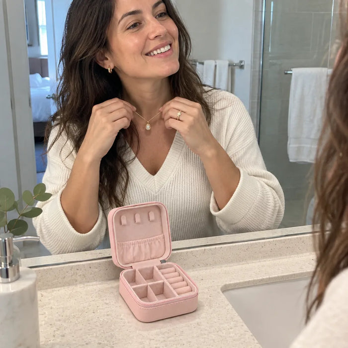 Smiling woman putting on a necklace at a hotel mirror, her travel jewelry case open on the counter