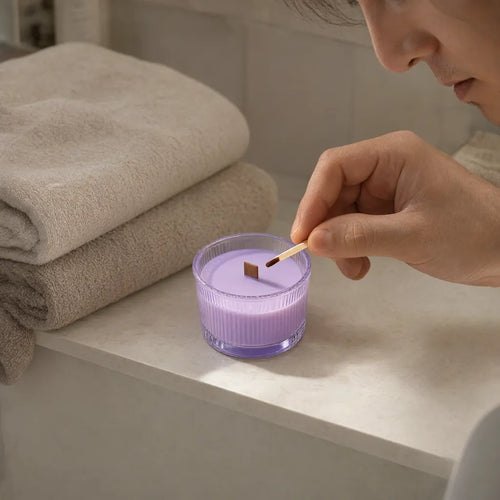 Man lighting a lavender scented candle on a bathroom shelf next to folded towels
