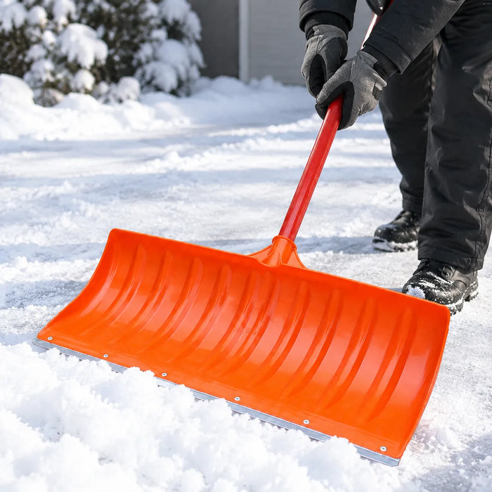 Hender i hansker holder skaftet på en oransje snøskuffe med bladet flatt på snøen