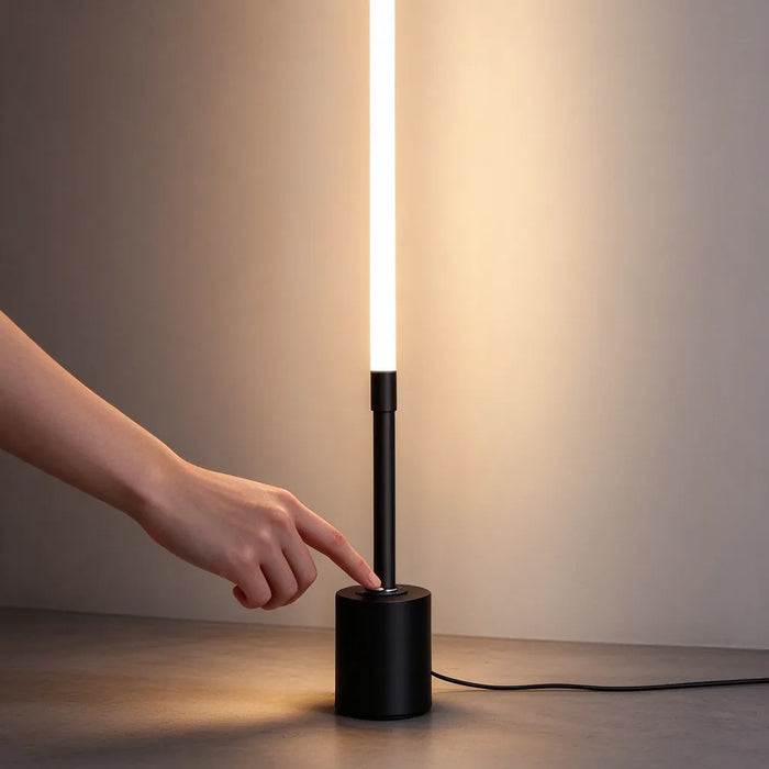 Finger switching on a slim tall LED floor lamp at the black base, warm light glowing