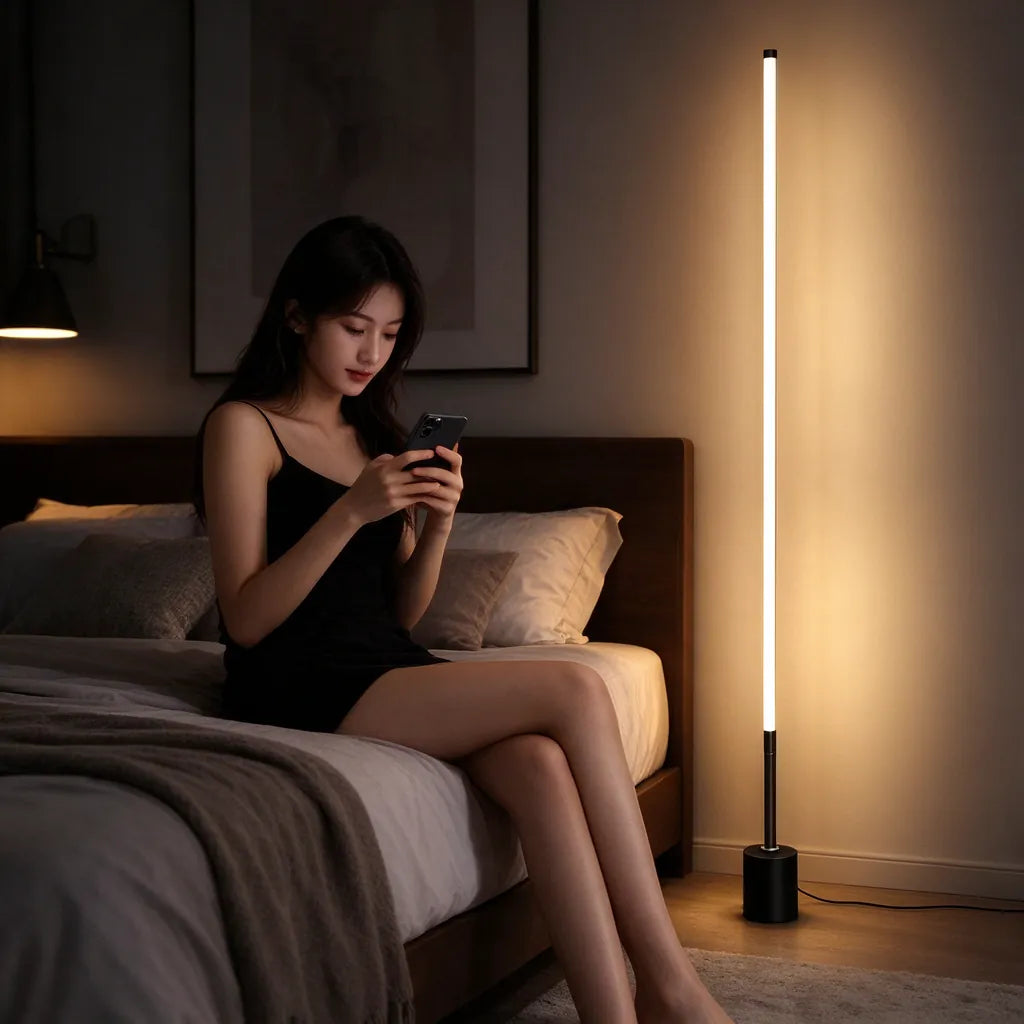 Woman on a bed with her phone beside a tall black floor lamp glowing warm in the bedroom