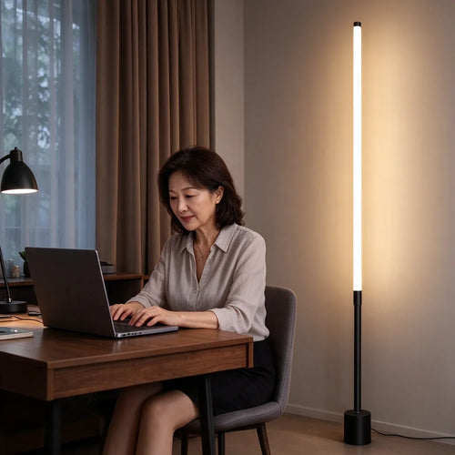 Woman working on a laptop at a home desk with a tall LED corner lamp glowing behind her