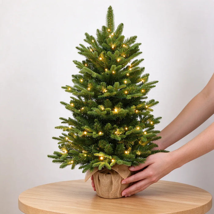 Hands placing a small artificial Christmas tree with lights on a wooden table