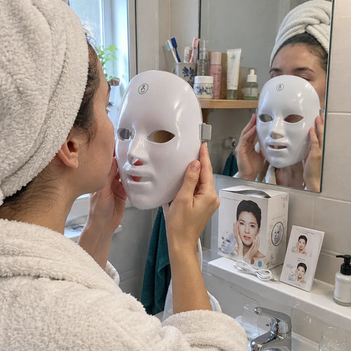 Junge Frau im Bademantel setzt am Lavabo eine LED Gesichtsmaske auf
