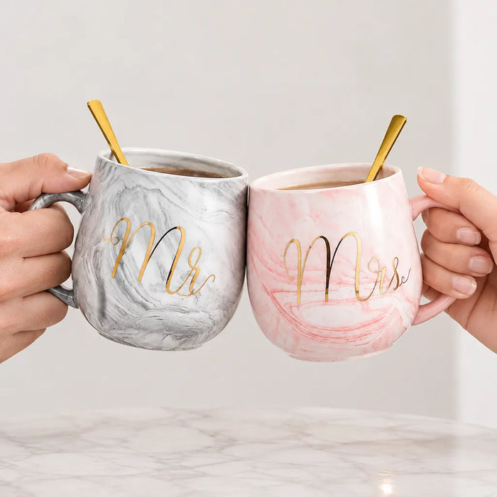 Two hands clinking his and hers marble mugs, grey Mr and pink Mrs, with gold spoons