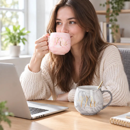 Woman sipping from a pink marble Mrs mug at her desk, grey Mr mug beside the laptop