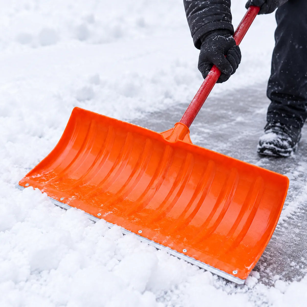 Gloved hands pushing snow with an orange snow shovel with a wide blade and red handle